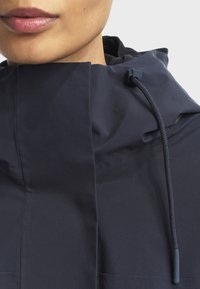 Mornarskoplavi kaput s visokim ovratnikom, detaljima na vezanje i glatkom teksturom. Ima elegantan dizajn i minimalistički izgled.