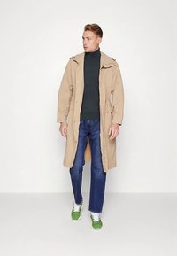 Beiger langer Mantel mit Kapuze und Kordelzug in der Taille, getragen über einem dunklen Rollkragenpullover, kombiniert mit blauen Jeans und grünen Sneakers.