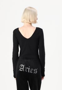 Svart langermet bodysuit med dyp V-bak. Kombinert med svarte fløyelsbukser med sølvfarget "Aries"-tekst i en stilisert font på baksiden.