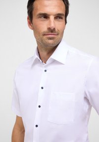 Korte mouwen witte blouse van een gladde stof, met blauwe knopen en een enkele borstzak. De kraag is gestructureerd en scherp.