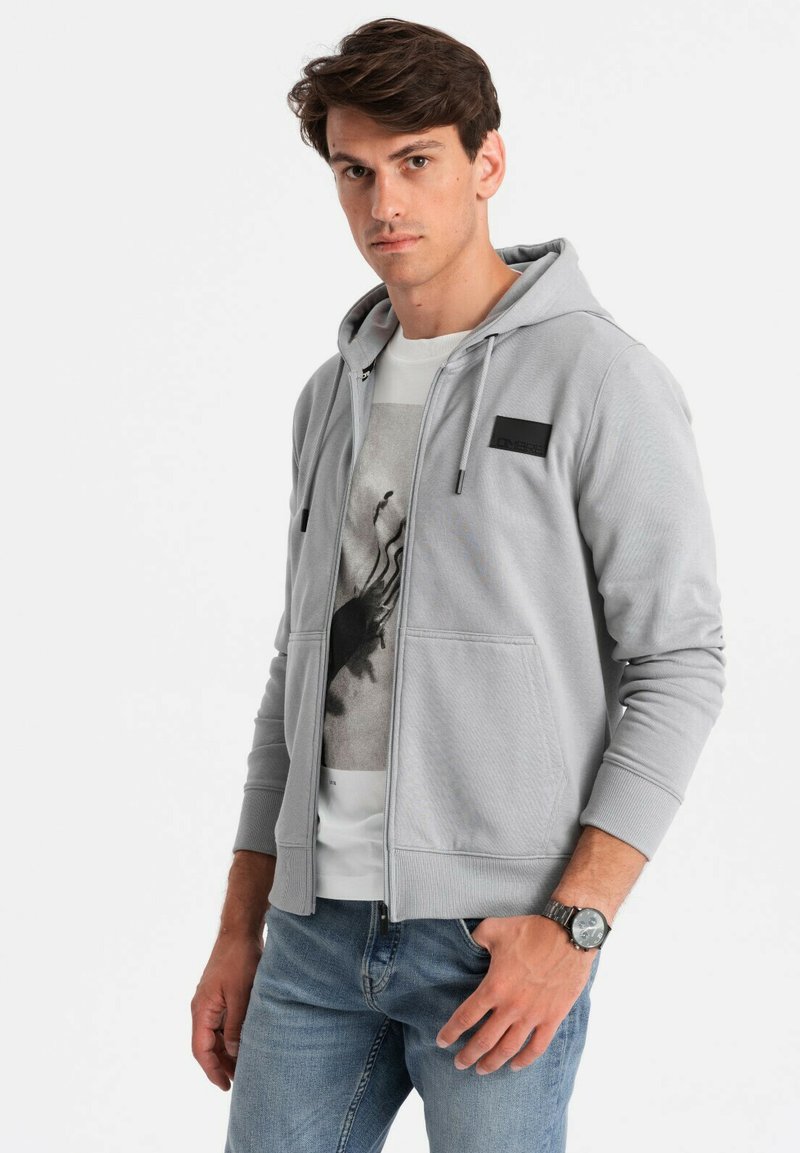 Sweat à zip gris avec un patch noir, tissu texturé, poches avant, porté sur un t-shirt blanc à motif, associé à un jean bleu.