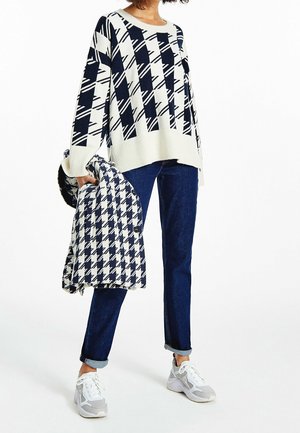 Pull en tricot à motifs bleu marine et blanc, ample avec des manches larges, associé à un jean en denim foncé et des baskets grises, tenant une veste assortie.