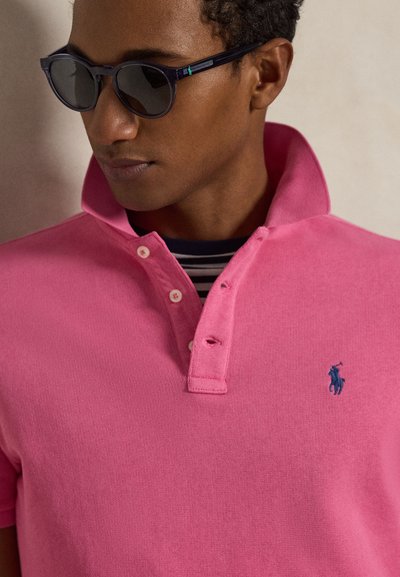 Άνδρας με σκούρα γυαλιά ηλίου, ροζ πόλο πουκάμισο με ανασηκωμένο γιακά και ριγέ πουκάμισο από κάτω, κοιτάζοντας προς τα κάτω και πλάγια.