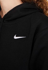 Person som bär en svart huvtröja med en vit Nike-swoosh-logotyp på vänster bröst, visar en del av sin nacke och lockigt hår.