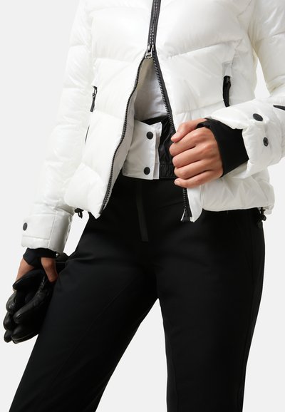 Witte pufferjack met zichtbare contrasterende stiksels, zwarte rits en knopen; gecombineerd met zwarte broek en zwarte handschoenen, wat een praktische uitstraling toont.