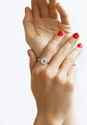 Silver ring with a circular design featuring a central hole, surrounded by small sparkling stones. Hands have red nail polish.