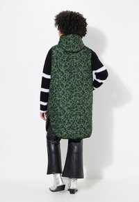 Gilet puffer con stampa leopardata verde e cappuccio, abbinato a un maglione a righe bianco e nero, pantaloni in eco-pelle nera e stivali metallici argento.