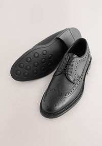 Chaussures habillées en cuir noir avec finition texturée, détails brogues et semelle en caoutchouc ronde présentant des motifs circulaires en relief.