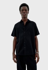 Junger Mann, der frontal steht, mit einem schwarzen strukturierten kurzärmligen Hemd und passenden Hosen, auf einem schlichten hellgrauen Hintergrund.