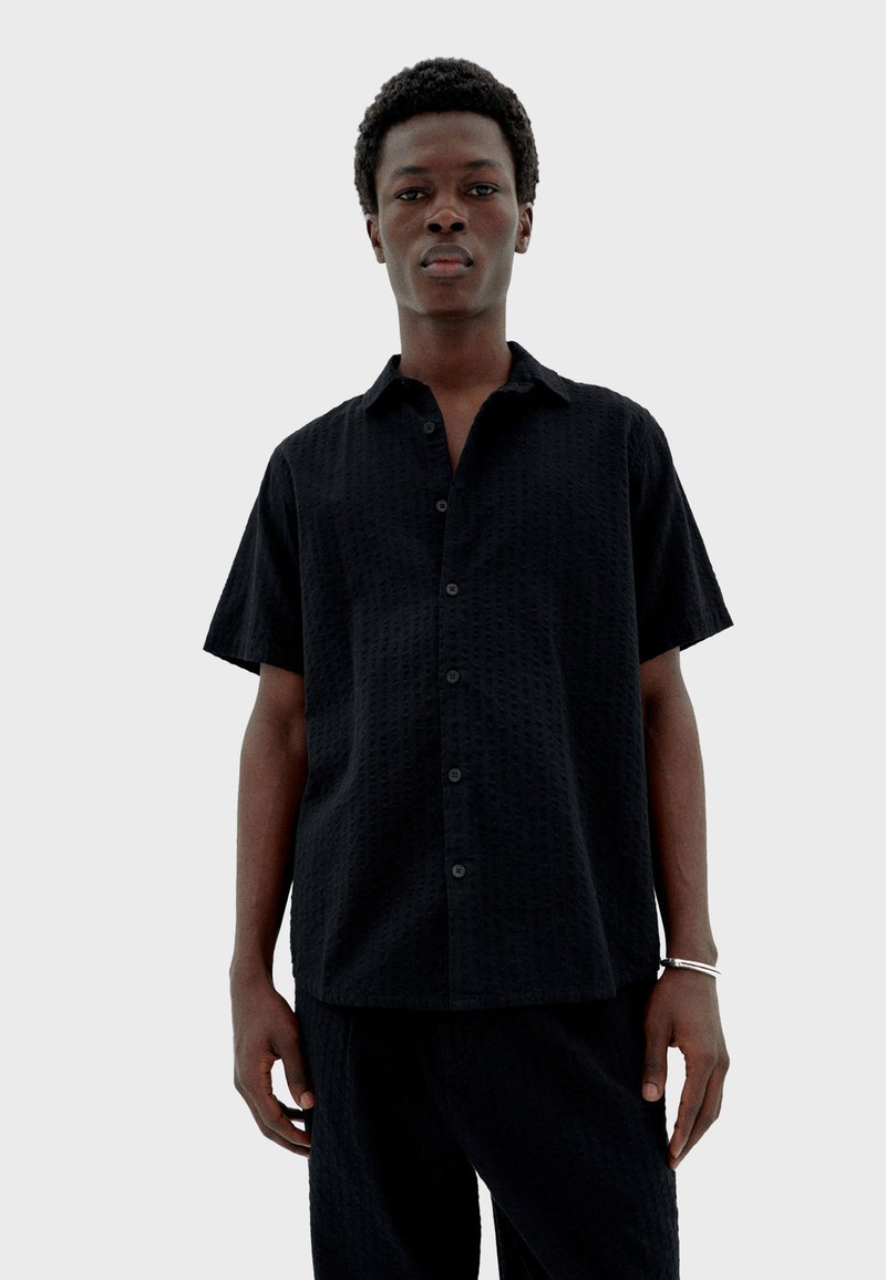 Junger Mann, der frontal steht, mit einem schwarzen strukturierten kurzärmligen Hemd und passenden Hosen, auf einem schlichten hellgrauen Hintergrund.