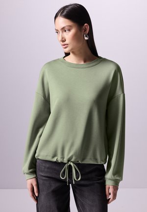 Grüne Sweatshirt mit lockerer Passform, langen Ärmeln, geripptem Ausschnitt und Kordelzugsaum. Kombiniert mit dunkelgrauen Hosen, glatte Textur.