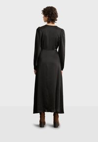 Vestido negro de mangas largas con un acabado suave y satinado. Presenta mangas abullonadas y una falda fluida. Combinado con botas de ante marrón hasta el tobillo.