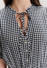 Camicia in gingham bianco e nero con scollatura arricciata, chiusura con fiocco, bottoni verticali e maniche corte a sbuffo indossata da una persona.