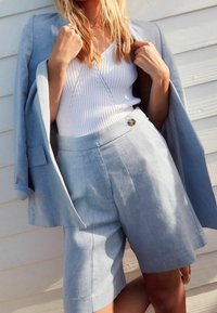 Blazer en lin bleu clair sur un débardeur blanc à côtes. Short bleu clair assorti avec une fermeture à bouton. Texture subtile et design ajusté.