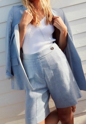 Lichtblauwe linnen blazer over een witte geribbelde tanktop. Bijpassende lichtblauwe shorts met een knoopsluiting. Subtiele textuur en tailormade ontwerp.