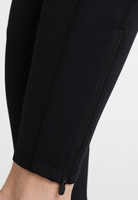 Černé legíny vyrobené z elastického materiálu, s kapsou na zip na kotníku a vertikálními švy podél nohavic pro zvýraznění detailů.