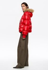 Czerwona puchowa kurtka z błyszczącym wykończeniem, kapturem wyściełanym futrem, elastycznymi mankietami i kieszeniami na zamek, w połączeniu z luźnymi oliwkowo-zielonymi spodniami.