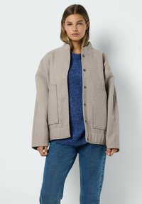 Veste en laine mélangée beige oversized avec un col montant, deux poches frontales et fermeture à boutons-pression ; portée sur un pull en maille bleu et un jean.