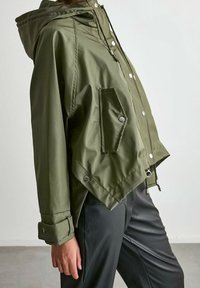 Olivgrön vattentät jacka med huva, tryckknappar, snedskärning och en framficka. Har elastisk dragsko i fållen.