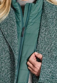 Gros plan sur la main d'une personne qui ferme une veste matelassée verte sous un manteau en laine texturée vert et blanc et un pull en tricot gris.