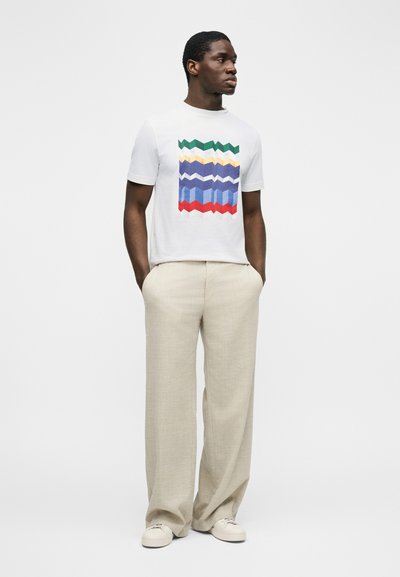 Άνδρας στέκεται με τα χέρια στις τσέπες, φοράει λευκό μπλουζάκι με πολύχρωμο γεωμετρικό σχέδιο, μπεζ παντελόνι φαρδιάς γραμμής και λευκά αθλητικά παπούτσια.