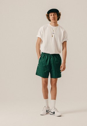 Junger Mann in weißem T-Shirt, grünen Nadelstreifen-Shorts, weißen Socken, weißen Turnschuhen, schwarzer Baskenmütze und goldenen Halsketten, der vor einem schlichten Hintergrund steht.