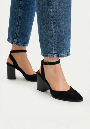 Stopy w czarnych zamszowych butach na blokowym obcasie z paskami wokół kostek, w połączeniu z niebieskimi przyciętymi jeansami.