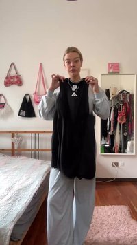 Black sleeveless top with a round neck and white logo detail, held by a person in a light-colored outfit. Room decor in the background.