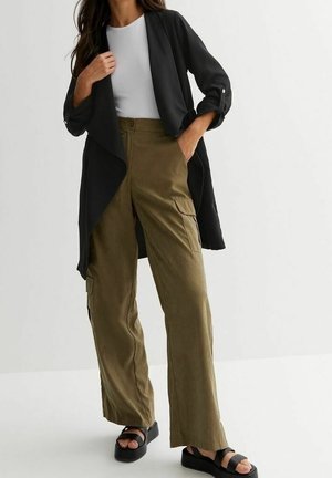 Femme portant un haut blanc, un cardigan noir ouvert devant, un pantalon cargo olive et des sandales à plateforme noires, debout avec une main dans la poche.