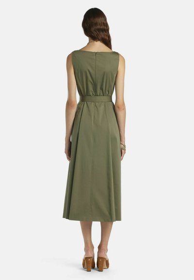 Sieviete ar gariem brūniem matiem, tērpusies bezroku olīvu zaļā vidēja garuma kleitā ar aizmugurējo rāvējslēdzēju, jostu viduklī un brūnu papēžu sandalēm, skatās prom.