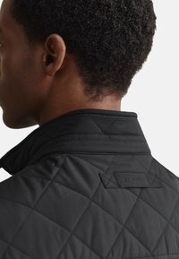 Sort quiltet jakke med høj krave og en lille logotag bagpå. Materialet har en glat, mat tekstur med diamantmønstre.