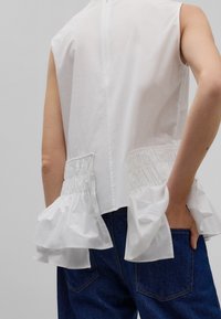 Top blanc à design sans manches, accents froncés sur les côtés et un dos légèrement allongé. Associé à un denim bleu.