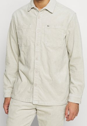 Man wearing light beige corduroy button-up shirt with two chest pockets and matching corduroy pants.
