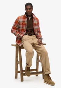Camisa a cuadros naranja y roja sobre un suéter marrón, combinada con pantalones beige y zapatos color canela, sentado en un taburete de madera. Look limpio y casual.