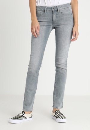 Schmal geschnittene hellgraue Jeans, getragen mit schwarz-weiß karrierten Slip-On-Sneakern, Hände locker an den Seiten, stehend vor einem schlichten hellen Hintergrund.