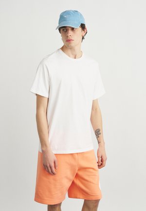 Nuori mies, joka käyttää valkoista t-paitaa, vaaleansinistä Nike-lippalakki, oransseja shortseja, seisoo vasemmassa kyynärvarressa olevan tatuoinnin kanssa yksivärisen harmaan taustan edessä.