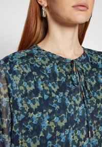 Femme portant des boucles d'oreilles en argent en forme d’anneaux et un chemisier foncé avec un motif floral bleu et vert, avec un col noué.