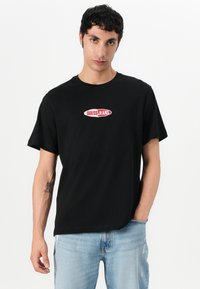 Black short-sleeve T-shirt featuring a central red and white "GUESS JEANS" logo on a curved badge. Casual fit, soft cotton texture.