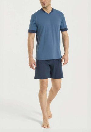 Uomo che cammina a piedi nudi indossando una maglietta blu con scollo a V e maniche corte con polsini scuri, e pantaloncini blu scuro su sfondo uniforme.