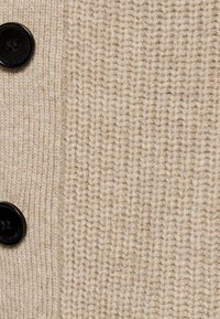 Tissu en laine tricotée beige avec texture côtelée et deux boutons noirs à quatre trous sur le côté gauche.