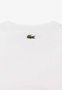 Camiseta blanca de algodón con cuello redondo, que presenta un pequeño logo de cocodrilo bordado en verde en la parte posterior. Textura suave y diseño sencillo.
