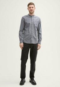 Camicia grigia con colletto a banda, maniche lunghe e tasca frontale, abbinata a pantaloni neri e scarpe, in piedi contro uno sfondo neutro.