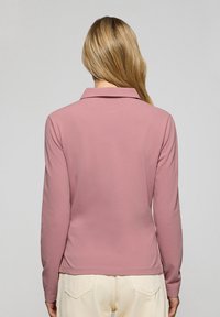 Långärmad skjorta med krage i ljusrosa tyg, med en slät textur och sidosprund vid fållen, bärs med krämfärgade byxor.