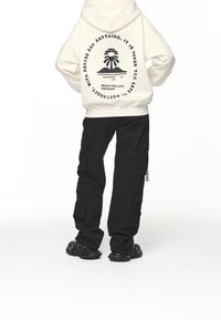 Persona che indossa un hoodie bianco con una grafica di palma e testo, pantaloni cargo neri e sneakers nere, in piedi su uno sfondo bianco.