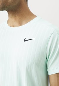 Mees, kes kannab heleda rohelise sooniku muster Nike spordipaita, millel on must Nike swoosh logo rinnal, lühendatud kaelast ja ülemisest käest.