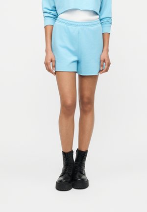 Person iført lyseblå shorts med elastik i taljen, matchende cropped sweatshirt og sorte snørestøvler med plateau, stående mod en ensfarvet baggrund.