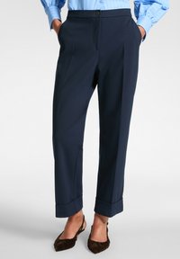 Marineblauwe broek met een getailleerde pasvorm, voorzien van een knoopsluiting, zijzakken en opgerolde manchetten. Gemaakt van een zachte stof met een subtiele glans.