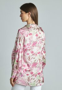 Blusa floreale in rosa, bianco e verde. Presenta maniche a palloncino, un colletto e dettagli con bottoni ai polsini. Tessuto leggero e liscio.