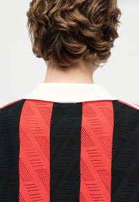 Κοντά καστανά κυματιστά μαλλιά που φαίνονται από πίσω, φορούν ένα κόκκινο και μαύρο πλεκτό πουκάμισο με κάθετες ρίγες και λευκό γιακά.