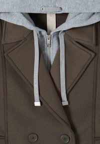 Manteau marron croisé avec revers, superposé sur un sweat à capuche gris avec une fermeture éclair, des cordons et le texte "DÉCOUVRIR DE NOUVEAUX ENDROITS".